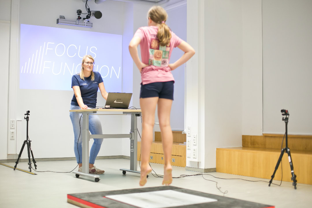 Schülerin führt Sprungtest auf Kraftmessplatte zur Return-to-Schulsport-Diagnostik durch, betreut von Sporttherapeutin im Trainingsraum